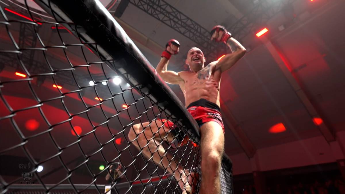 Zawodnik MMA z podniesionymi rękami w geście triumfu, stojący w klatce.