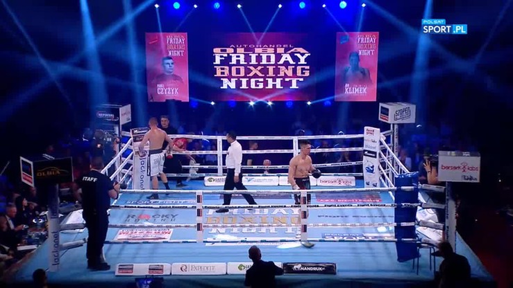 Scena z gali bokserskiej z zawodnikami w ringu, sędzią i telebimami z logotypem "Olba Friday Boxing Night".