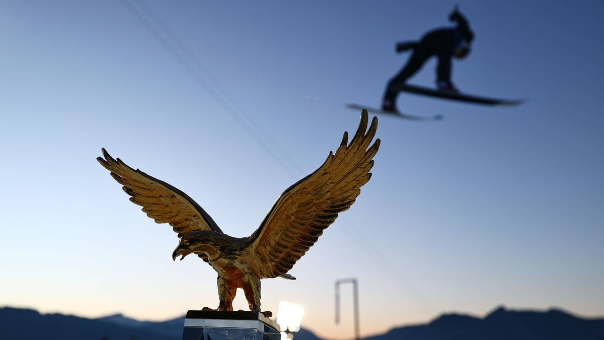 Złota statua orła w pierwszym planie, skoczek narciarski w locie na dalszym planie.