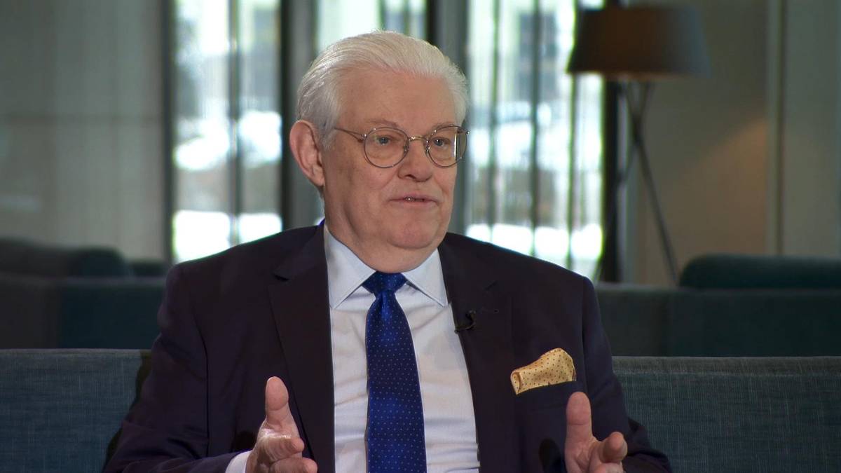 Starszy mężczyzna w garniturze i krawacie siedzi i m&oacute;wi.