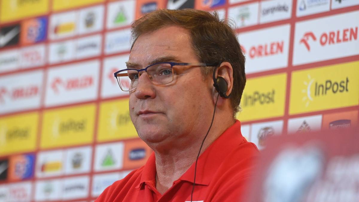 Trener Jan Urban w czerwonej koszulce z orłem na piersi, w okularach, podczas konferencji prasowej.