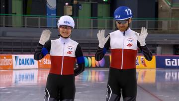 Dwóch łyżwiarzy szybkich w strojach z polskimi barwami narodowymi, z białymi kaskami i białymi rękawiczkami, pozuje do zdjęcia na lodowisku.