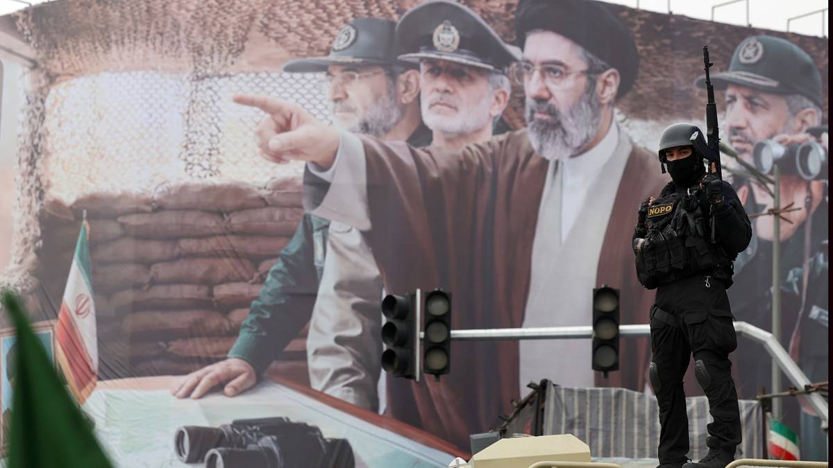 Żołnierz w czarnym stroju i hełmie stoi przed wielkim muralem z wizerunkami irańskich przyw&oacute;dc&oacute;w.