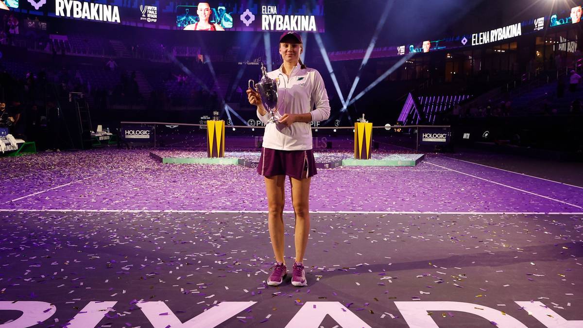 Elena Rybakina trzyma puchar na korcie tenisowym, w tle podświetlane trybuny.