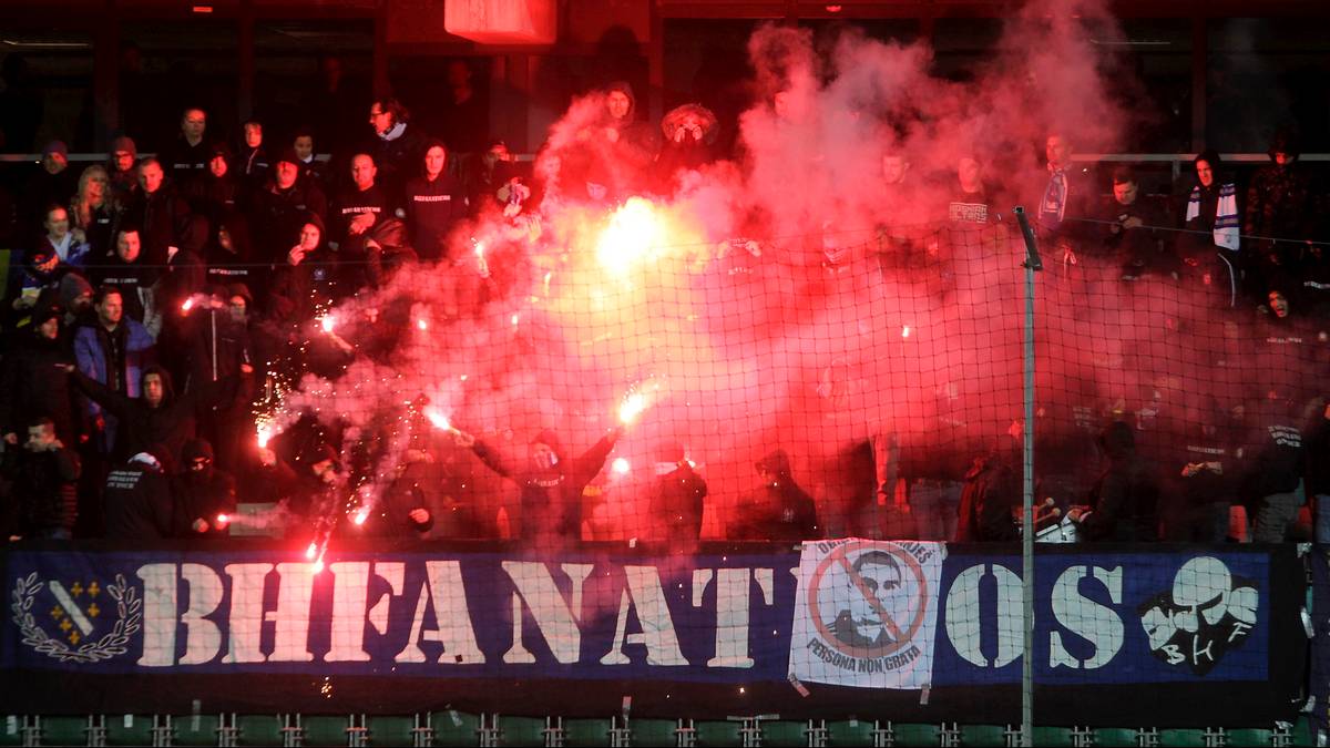 Transparent "BH FANATICOS" na trybunie stadionu podczas meczu, z kibicami i czerwonymi racami.
