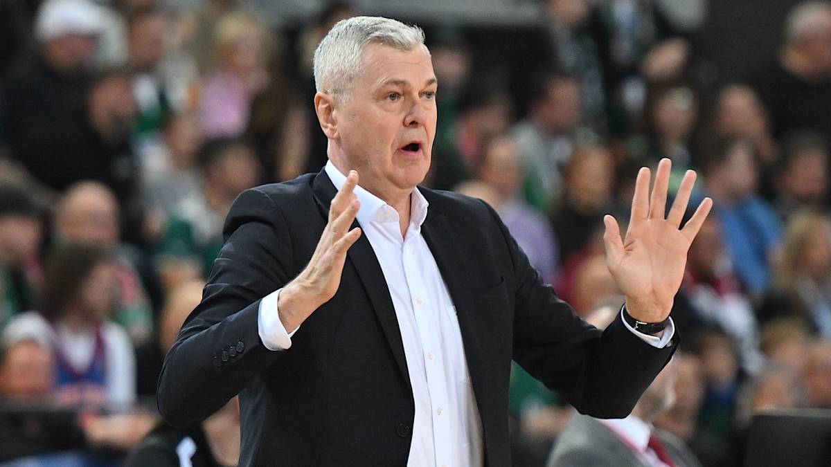 Trener w czarnym garniturze i białej koszuli, z siwymi włosami, gestykuluje rękami podczas meczu koszykówki. W tle widać publiczność.