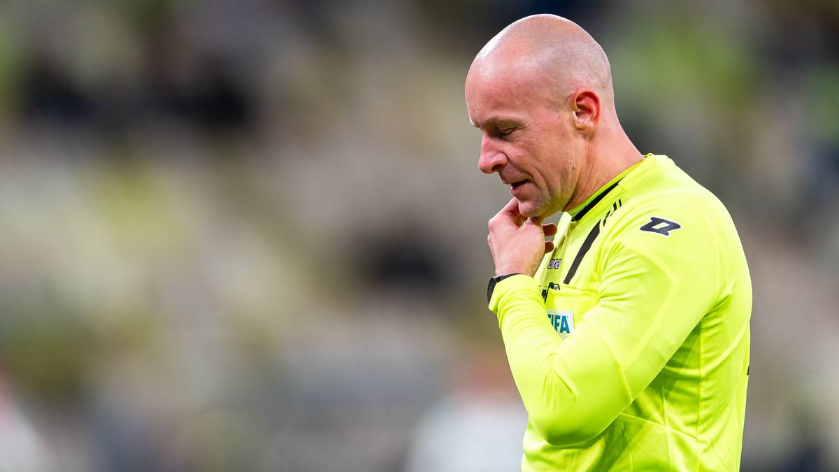 Szymon Marciniak, sędzia piłkarski, w żółtej koszulce z długim rękawem, patrzy w bok z zamyśloną miną, opierając dłoń na podbródku.