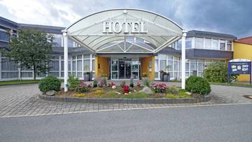 Wejście do nowoczesnego hotelu z dużym zadaszeniem i ozdobnym klombem przed drzwiami.