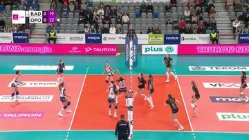 Mecz siatk&oacute;wki kobiet pomiędzy Moya Radomka Radom a UNI Opole. Zawodniczki w akcji przy siatce, jedna z nich pr&oacute;buje wykonać atak, podczas gdy przeciwniczki przygotowują się do bloku. Na trybunach widoczni są kibice.