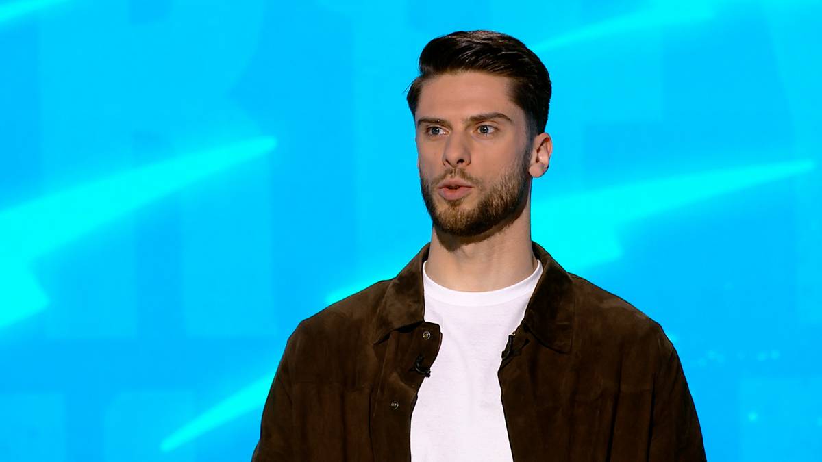 Młody mężczyzna z brodą, ubrany w brązową kurtkę i białą koszulkę, patrzy przed siebie na jasnoniebieskim tle.