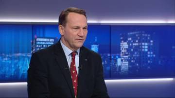 Radosław Sikorski w garniturze przed panelem świetlnym, z nocnym miastem w tle.