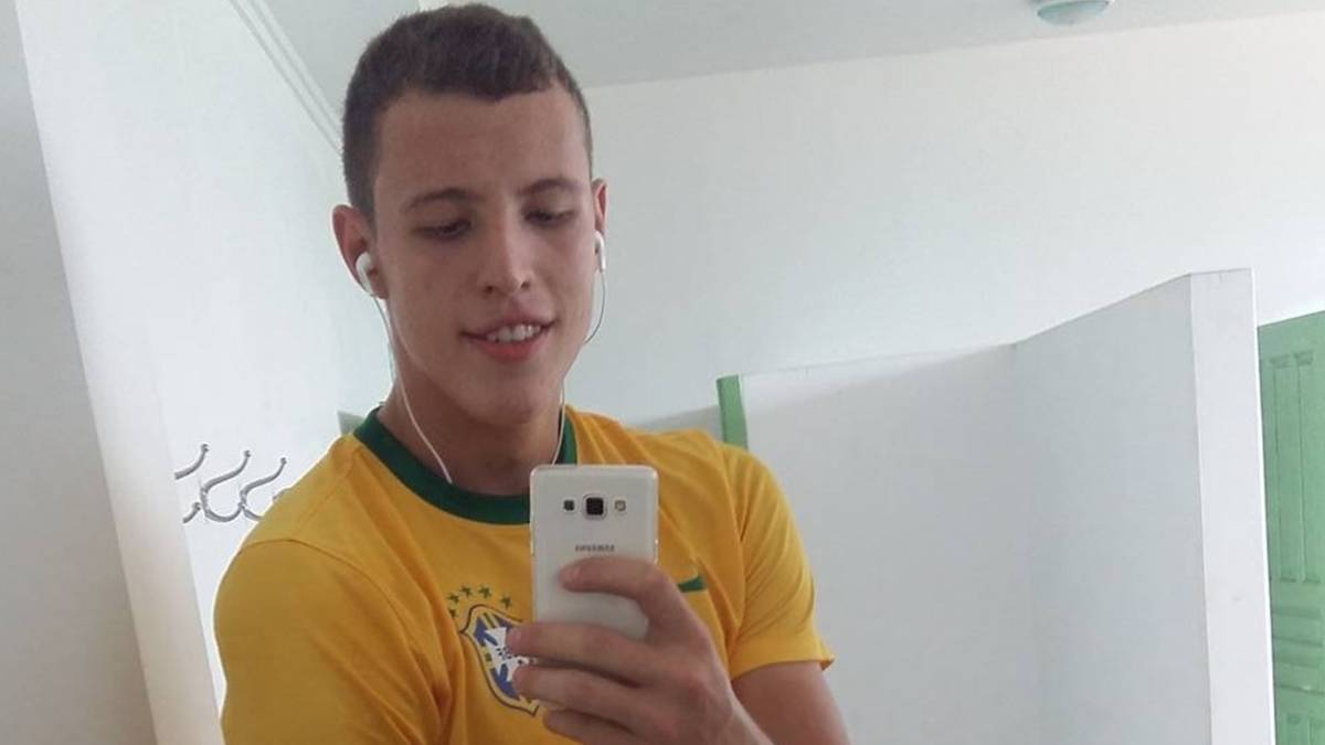 Młody mężczyzna w żółtej koszulce z zielonym kołnierzykiem, trzymający smartfon i słuchawki.