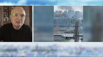 Starszy mężczyzna w okularach i czarnym swetrze, obok widok na miasto z unoszącym się dymem nad budynkami.