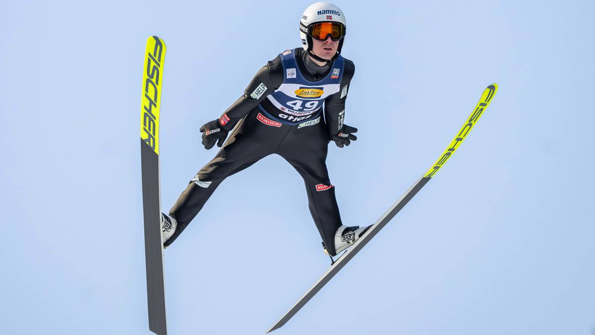 Skoczek narciarski w kombinezonie i kasku z goglami leci w powietrzu na nartach.