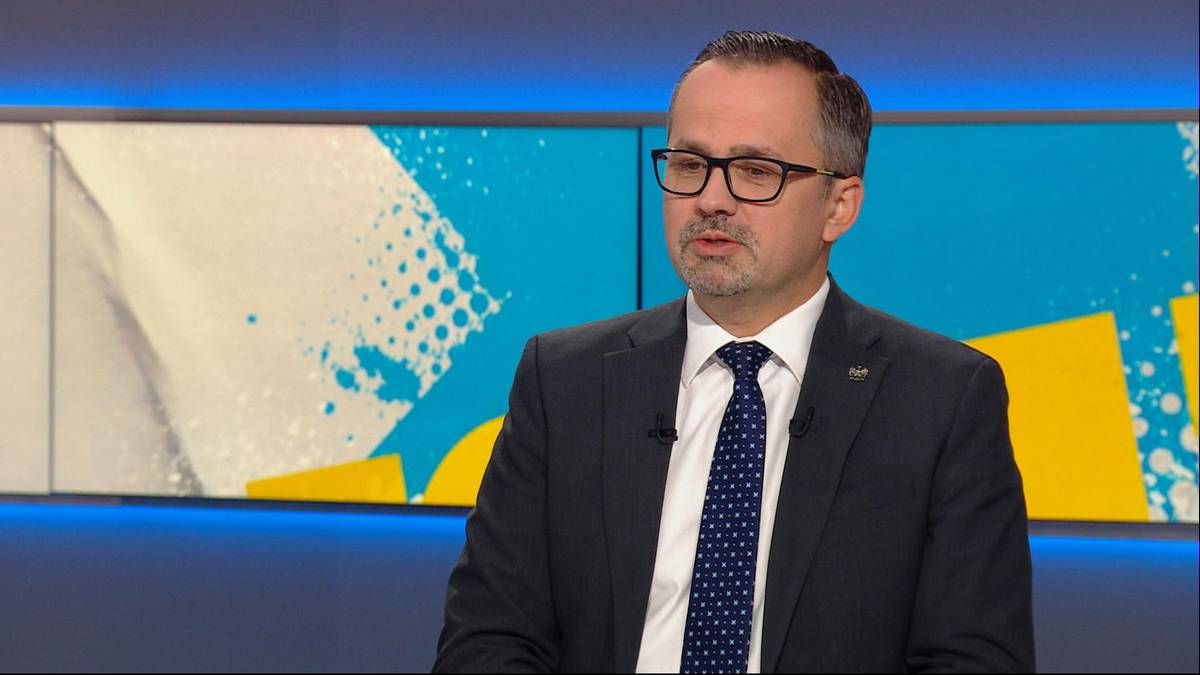 Mężczyzna w garniturze i okularach wypowiada się w studio telewizyjnym na tle niebiesko-ż&oacute;łtej scenografii.
