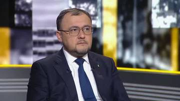 Mężczyzna w okularach i garniturze siedzi w studiu telewizyjnym.