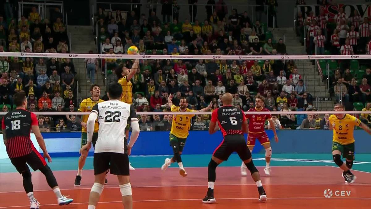 Siatkarze podczas meczu, jeden z nich wykonuje zagrywkę nad siatką, w tle widzowie na trybunach.