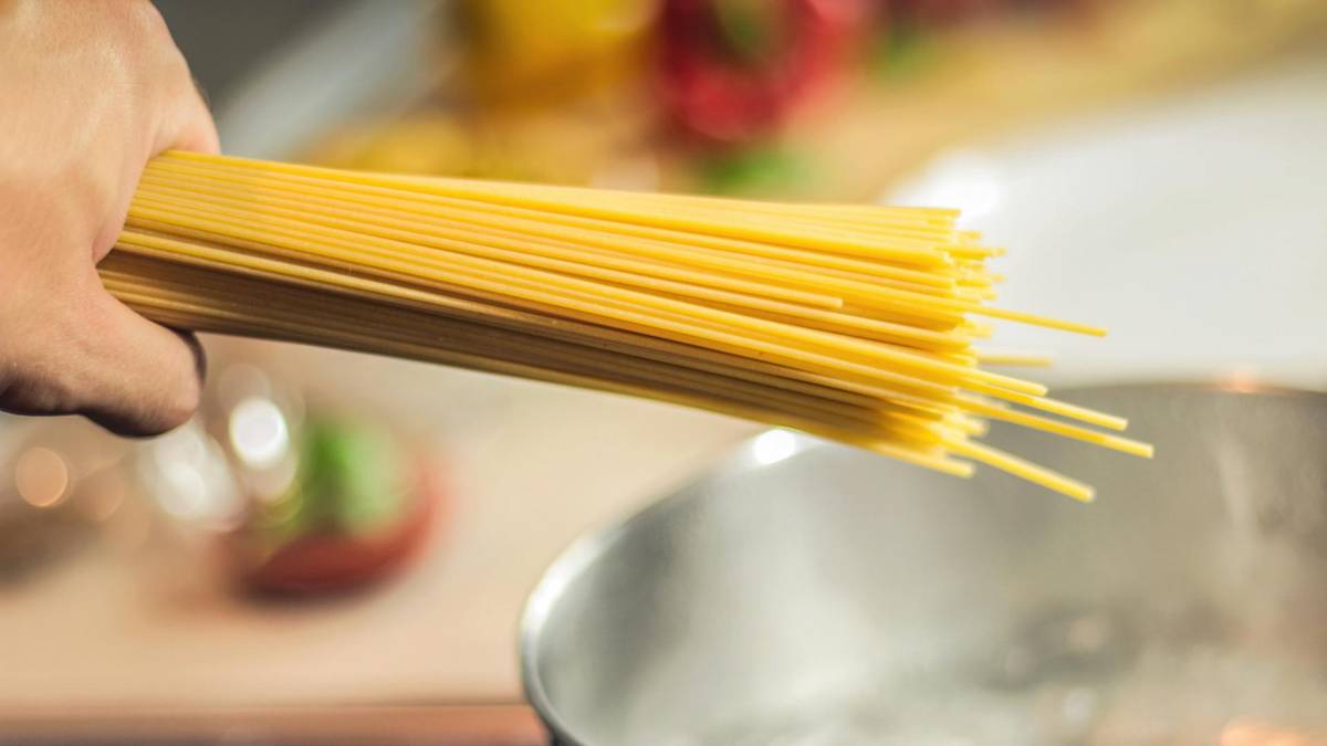 Dłoń osoby trzymająca wiązkę surowego makaronu spaghetti nad garnkiem z bulgoczącą wodą.