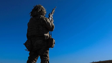 Żołnierz w umundurowaniu kamuflażowym trzyma karabin na tle błękitnego nieba.