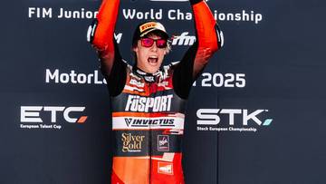 Młody kierowca wyścigowy w stroju z napisem "FUSPORT" i "INVVICTUS" triumfuje, unosząc ręce w górę.