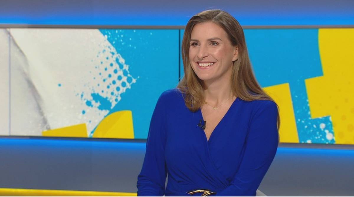 Kobieta w niebieskiej sukience siedzi na tle studia telewizyjnego z uśmiechem na twarzy.