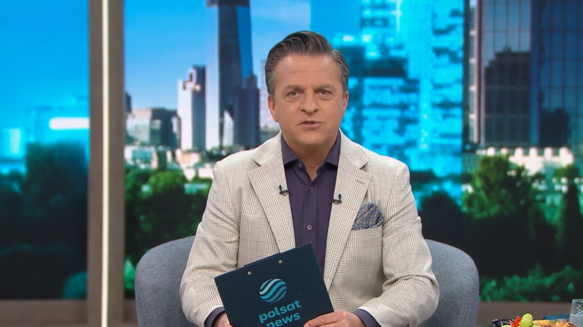 Mężczyzna w marynarce i koszuli siedzi w studiu telewizyjnym na tle panoramy miasta, trzymając w rękach podkładkę z logo Polsat News.