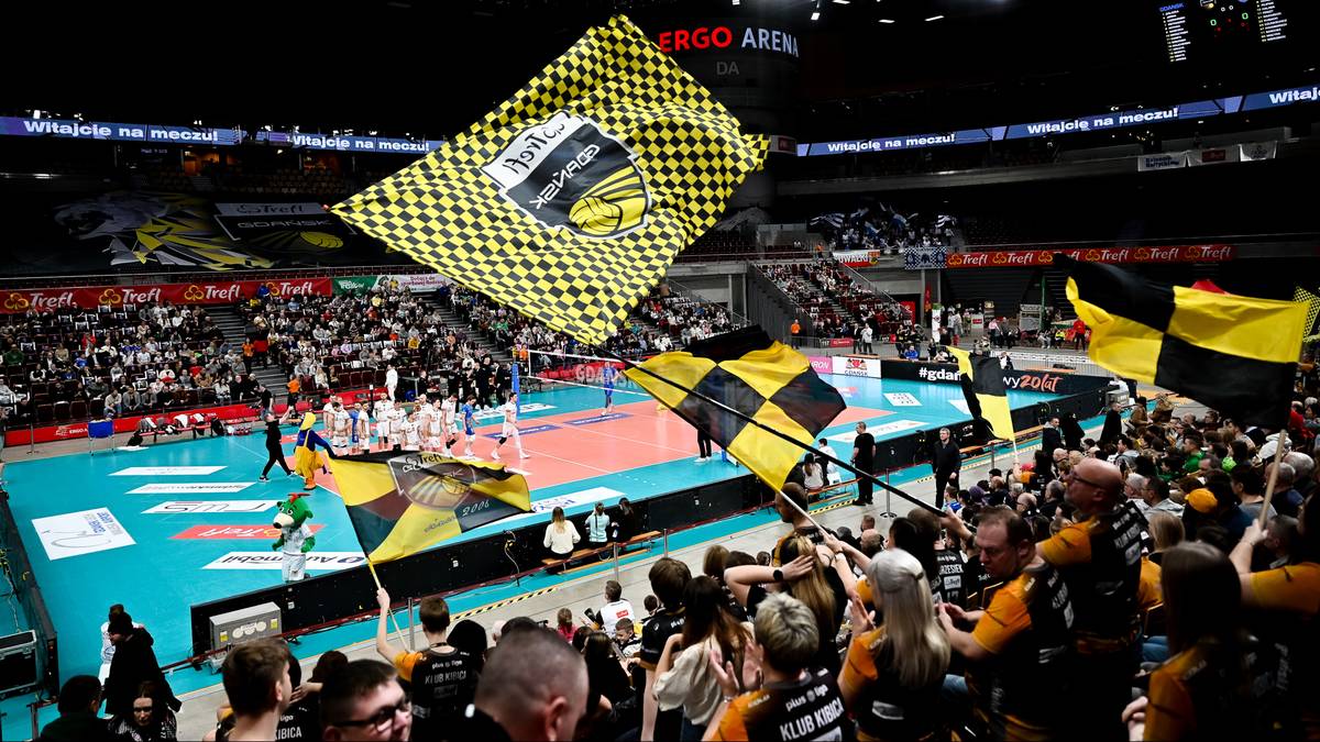 Widok z trybun na boisko siatkarskie podczas meczu, z kibicami machającymi ż&oacute;łto-czarnymi flagami w kratkę.