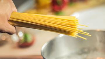 Dłoń osoby trzymająca wiązkę surowego makaronu spaghetti nad garnkiem z bulgoczącą wodą.