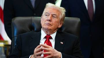 Donald Trump siedzący za biurkiem w garniturze, z dłońmi złożonymi przed sobą.