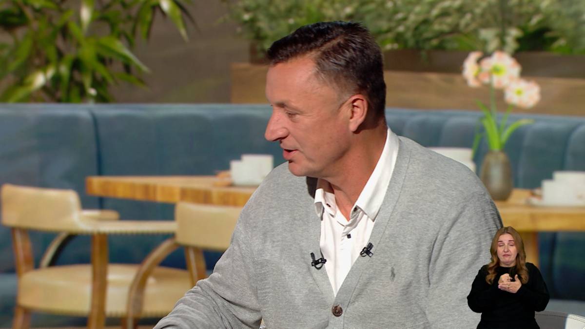 Tomasz Hajto siedzi i mówi, ubrany w szary sweter. W tle widać stoły i siedzenia, a w prawym dolnym rogu ekranu znajduje się tłumaczka języka migowego.