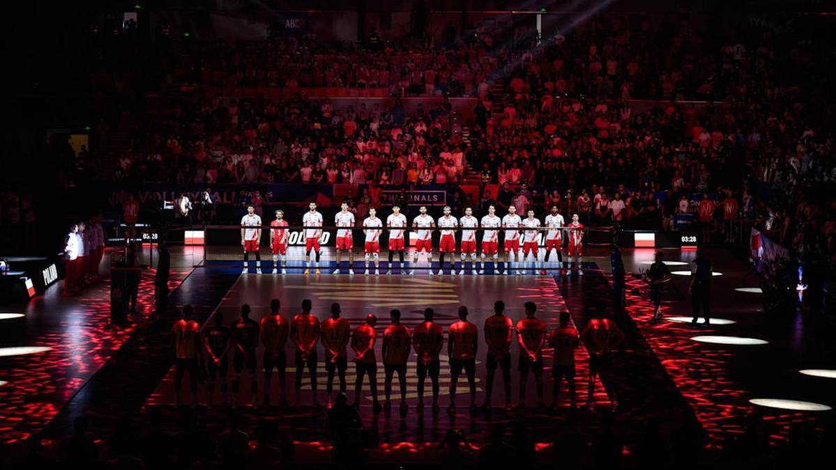 Drużyna siatkarzy w białych strojach, stojąca w dwóch rzędach na boisku oświetlonym czerwonymi światłami, przed wypełnioną po brzegi widownią.