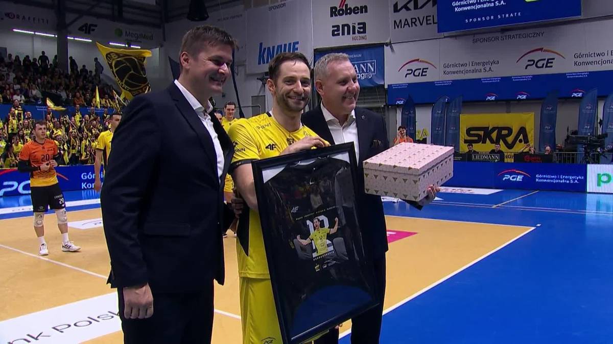 Dw&oacute;ch mężczyzn w ciemnych garniturach wręcza ramę z koszulką i ozdobne pudełko siatkarzowi w ż&oacute;łtej koszulce sportowej. Scena ma miejsce w hali sportowej, na tle kt&oacute;rej widać trybuny z kibicami.