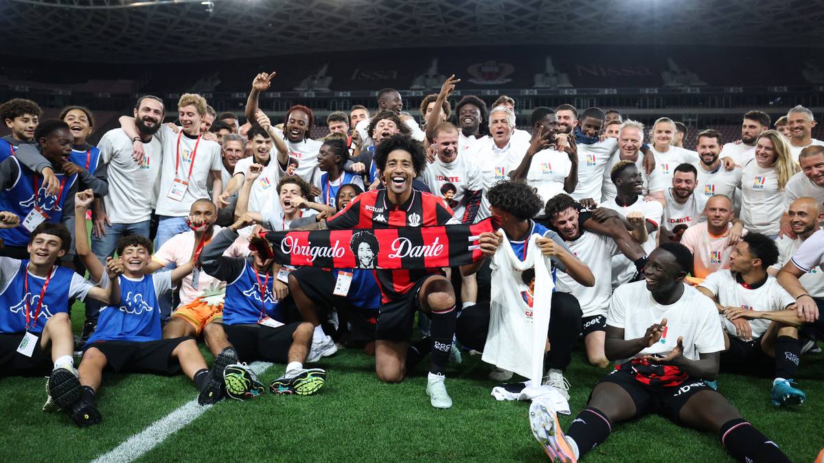 Grupa ludzi w strojach sportowych, prawdopodobnie drużyna piłkarska, świętuje na zielonej murawie stadionu. Wszyscy uśmiechają się i pozują do zdjęcia. Na pierwszym planie jeden z nich trzyma transparent z napisem "Danke".