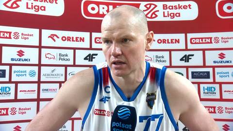 Damian Kulig, koszykarz z ogoloną głową, w stroju sportowym z numerem 77, udziela wywiadu z mikrofonem przed twarzą, na tle ściany z logotypami sponsor&oacute;w.