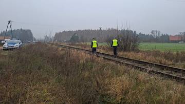 Dwóch policjantów w żółtych kamizelkach idzie wzdłuż torów kolejowych. W tle widać samochód policyjny i drzewa.