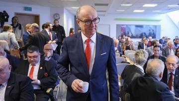 Mężczyzna w garniturze i okularach trzyma biały kubek, idąc przez salę pełną ludzi.