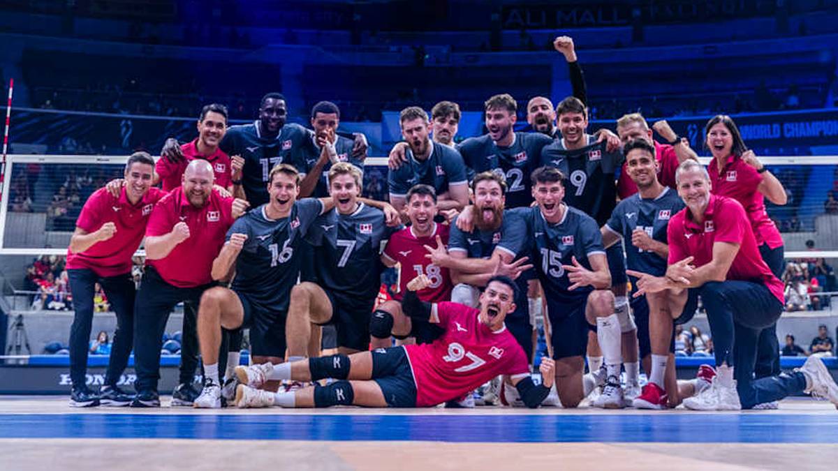 Drużyna siatkarzy w czerwonych koszulkach z numerami i białymi elementami, ubrani w niebieskie spodenki, pozuje do zdjęcia na boisku siatkarskim po wygranej. Gracze śmieją się, obejmują i świętują.