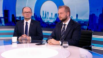 Dw&oacute;ch mężczyzn w garniturach siedzi przy okrągłym stole. Na stole znajdują się szklanki z wodą i telefon.