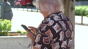 Starsza kobieta w koszuli w koty trzyma telefon w ręku.