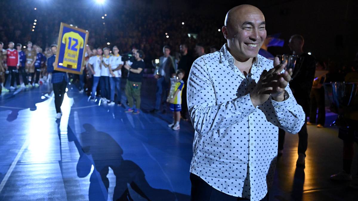 Uśmiechnięty łysy mężczyzna w koszulce w kropki klaszcze w dłonie na tle tłumu i hali sportowej.
