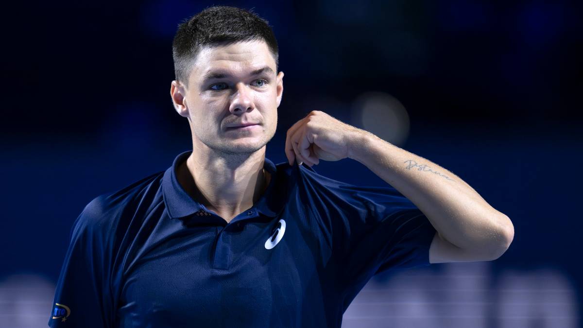 Młody mężczyzna w ciemnoniebieskiej koszulce polo z białym logo, poprawia kołnierzyk, patrząc w bok.