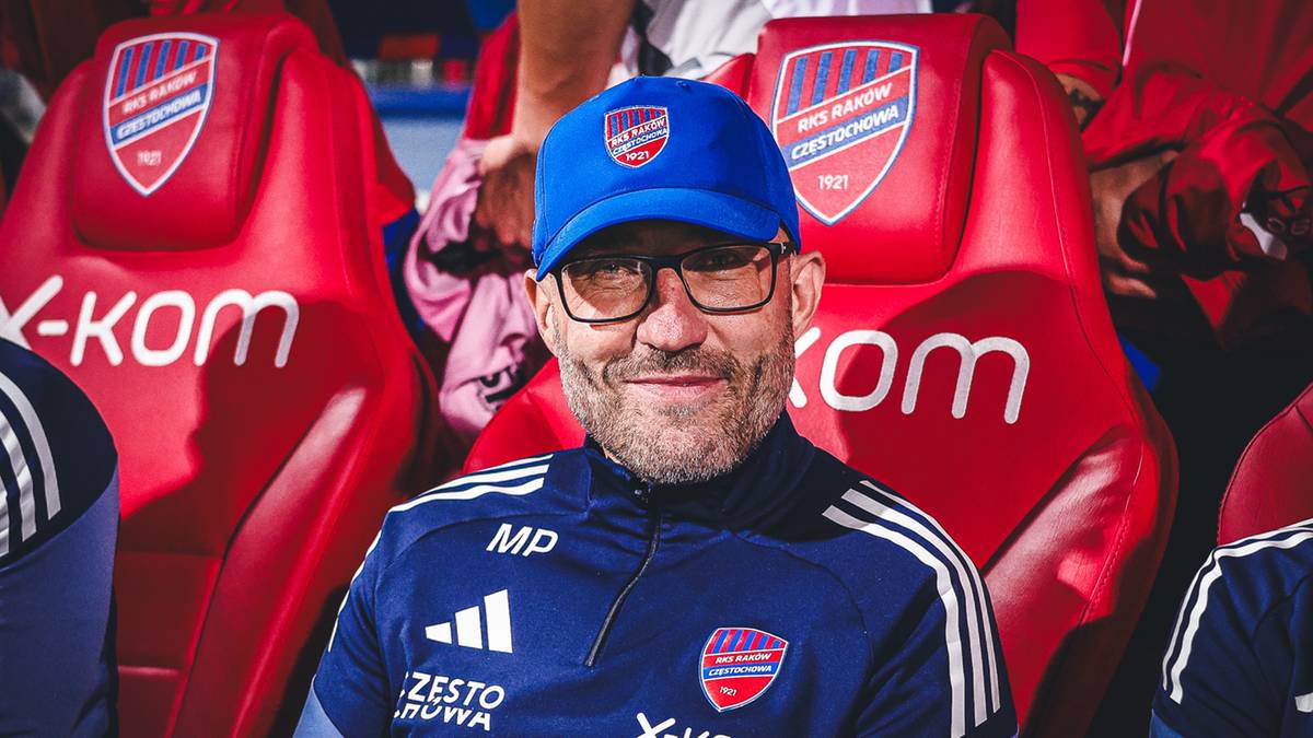 Uśmiechnięty mężczyzna w niebieskiej czapce z daszkiem i okularach siedzi na tle czerwonych siedzeń z logo drużyny.