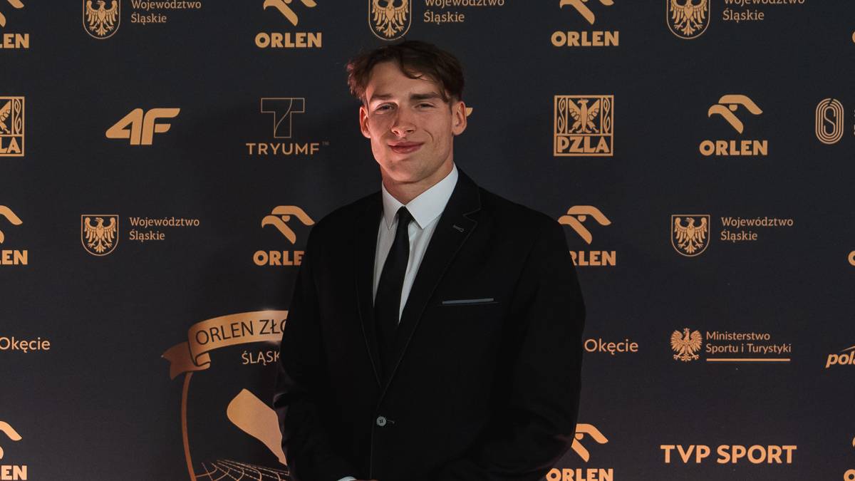 Młody mężczyzna w czarnym garniturze i białej koszuli z czarnym krawatem, stojący na tle ciemnej ściany ozdobionej złotymi logotypami sponsor&oacute;w i herbami. Mężczyzna patrzy na wprost i uśmiecha się lekko.