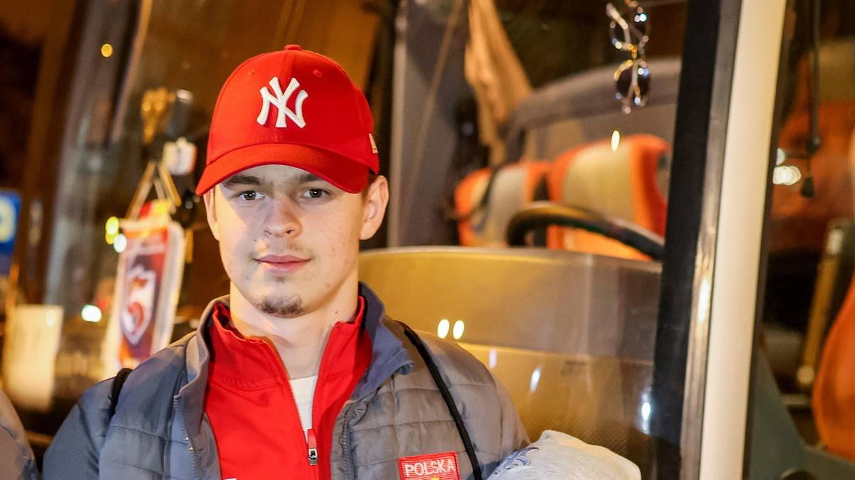 Młody mężczyzna w czerwonej czapce z daszkiem z logo NY i szarej kurtce, patrzący w obiektyw.