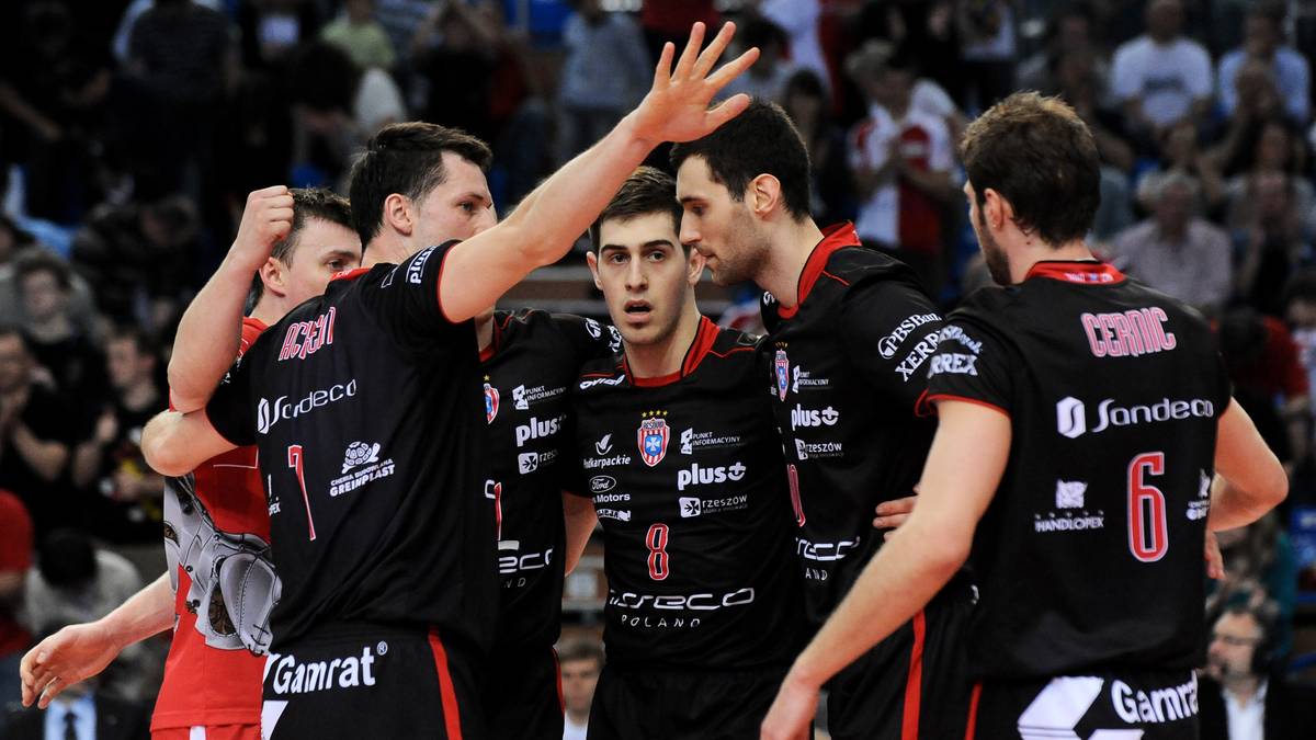 Grupa siatkarzy w czarnych koszulkach z czerwonymi elementami obejmuje się podczas przerwy w meczu.