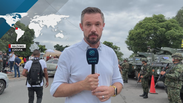 Korespondent Polsat News z mikrofonem stoi na tle uzbrojonych żołnierzy i pojazd&oacute;w wojskowych, w tle mapa świata z zaznaczonym regionem Ameryki Południowej.