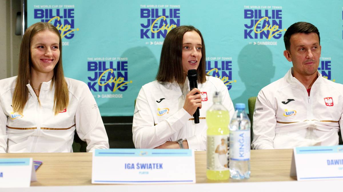 Trzy osoby siedzące przy stole, w tym Iga Świątek z mikrofonem, podczas konferencji prasowej Billie Jean King Cup.