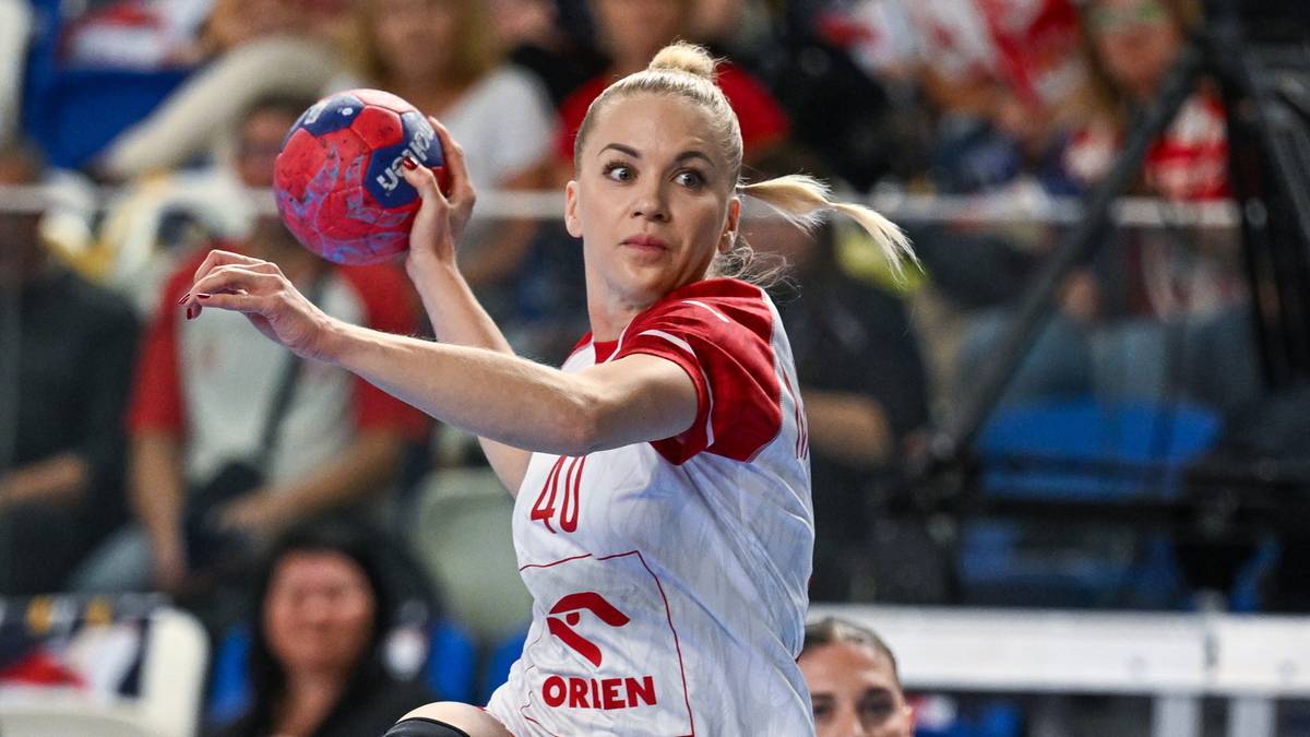 Piłkarka ręczna w białej koszulce z czerwonymi rękawami i numerem 40, trzymająca piłkę do piłki ręcznej w prawej dłoni, gotowa do rzutu. Tło jest nieostre, z widownią na trybunach.