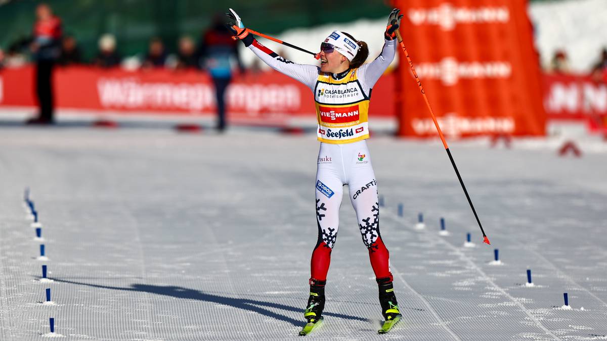 Zawodniczka narciarstwa biegowego z rękami uniesionymi w geście triumfu, trzymająca kije narciarskie.