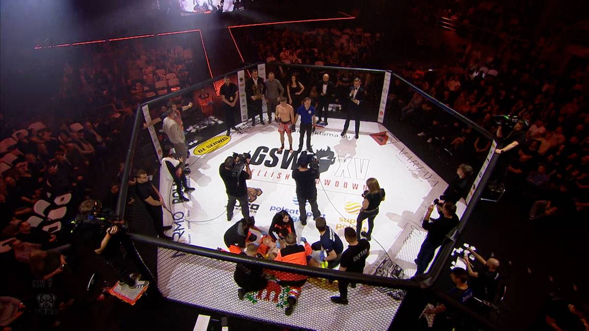 Widok z góry na arenę gali sportów walki w Polsce. W centralnej części znajduje się oktagonalna klatka, gdzie na noszach jest wynoszony zawodnik. Wokół klatki zgromadzeni są ludzie i personel.